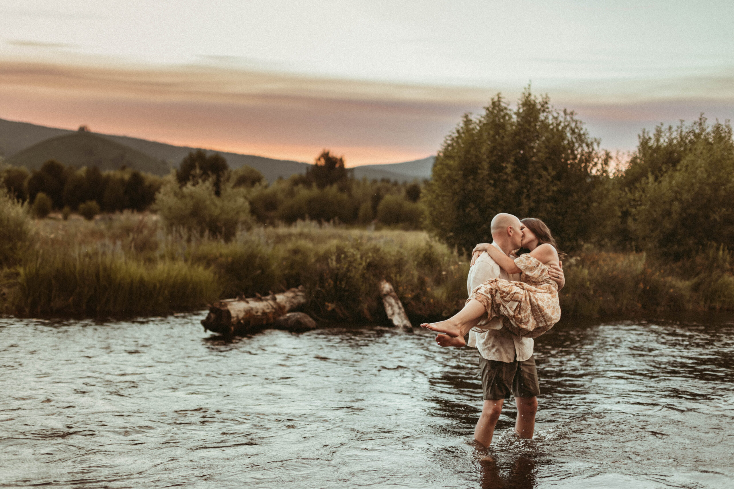 Man holding woman in arms in water while the sun sets